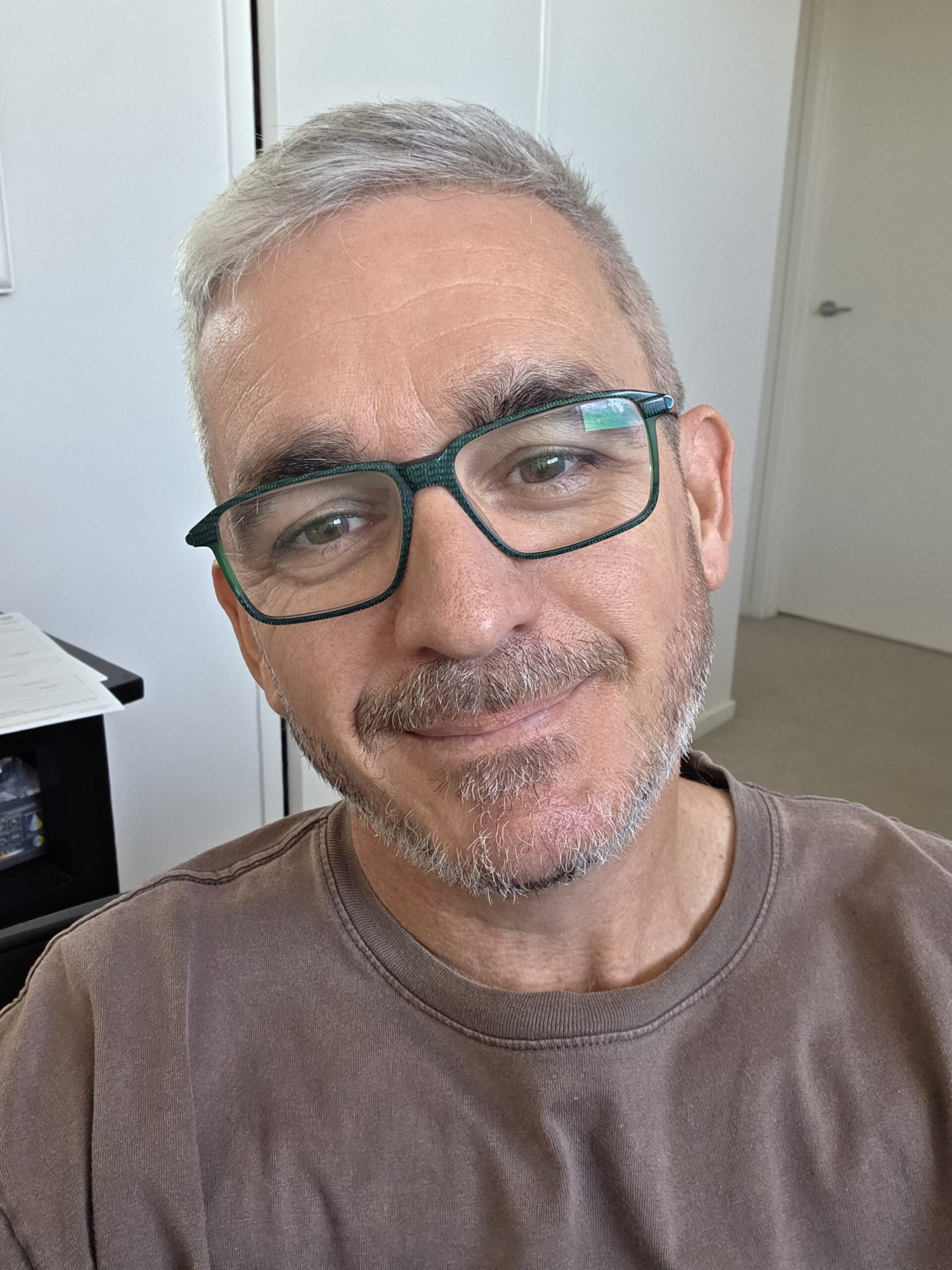A person with short grey hair, glasses, and a trimmed beard is smiling at the camera. An independent masseuse in a brown shirt sits indoors, with a white wall and door in the background.