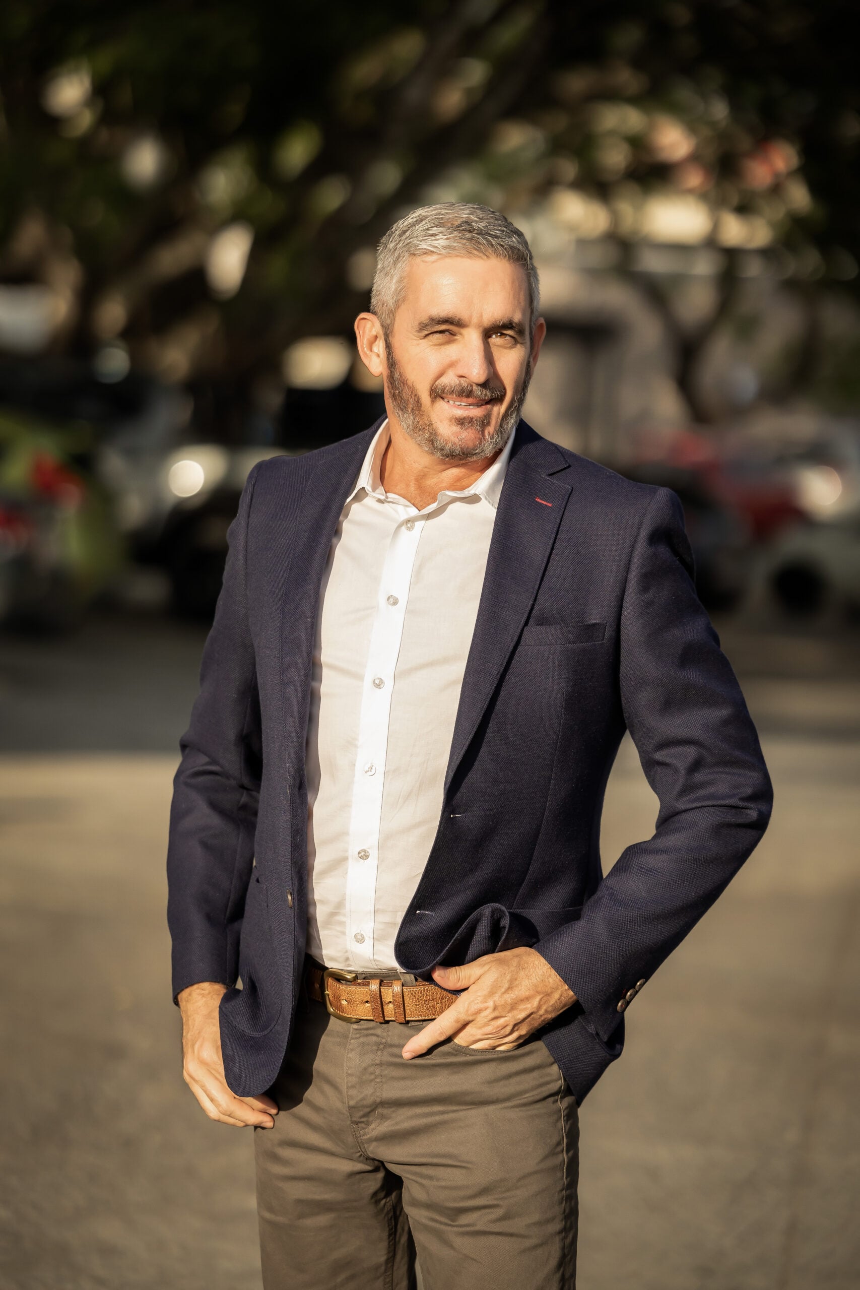 A man with short grey hair and a beard, wearing a navy blazer, white shirt, and brown belt, stands outdoors on a sunny day, smiling with one hand on his hip. The backdrop hints at relaxation found in sensual massage getaways.